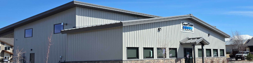 A modern, light gray metal building with a stone base and a dark gray roof. The building has several rectangular windows and a prominent sign with blue lettering under a small awning above the entrance.
